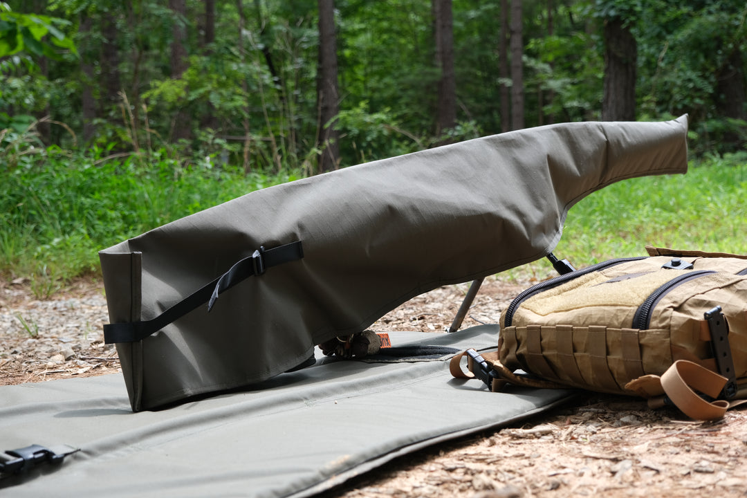 Suppressor Ready Rifle Cover protecting a precision rifle on a shooting mat in a wooded outdoor setting.