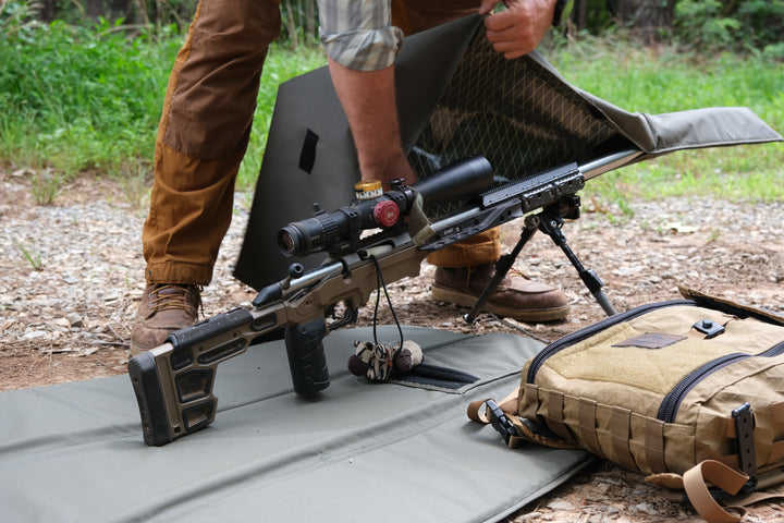 Precision rifle being uncovered from a Suppressor Ready Rifle Cover on a shooting mat in a wooded outdoor setting.