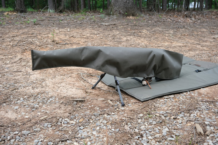 Suppressor Ready Rifle Cover protecting a precision rifle on a shooting mat in a wooded outdoor setting.